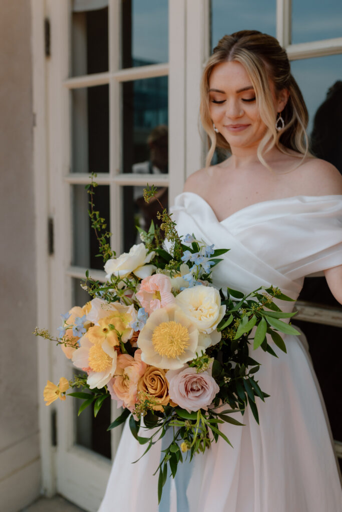 Close-up of bride holding a colorful bouquet with roses and wildflowers, wearing a classic off-shoulder wedding gown.