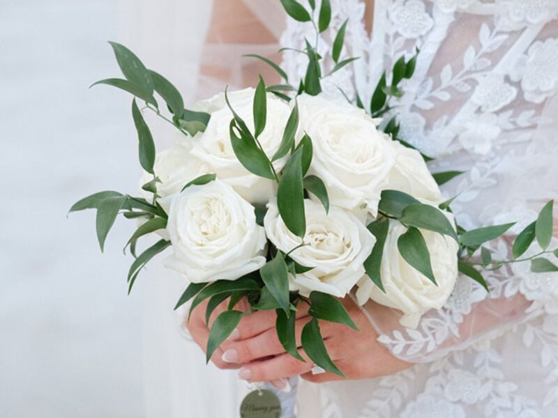 Close-up of a white rose bouquet accented with green leaves, held by a bride in a lace wedding dress.