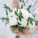 Close-up of a white rose bouquet accented with green leaves, held by a bride in a lace wedding dress.