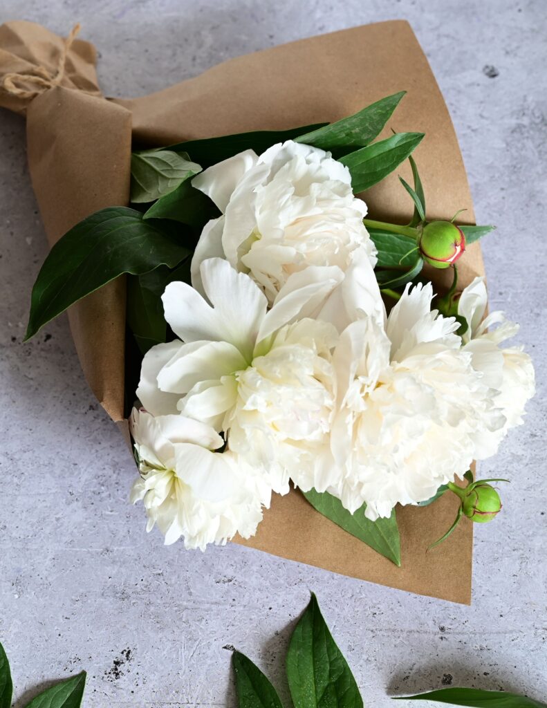 White peonies wrapped in brown paper with green leaves, creating a soft and rustic bouquet design.