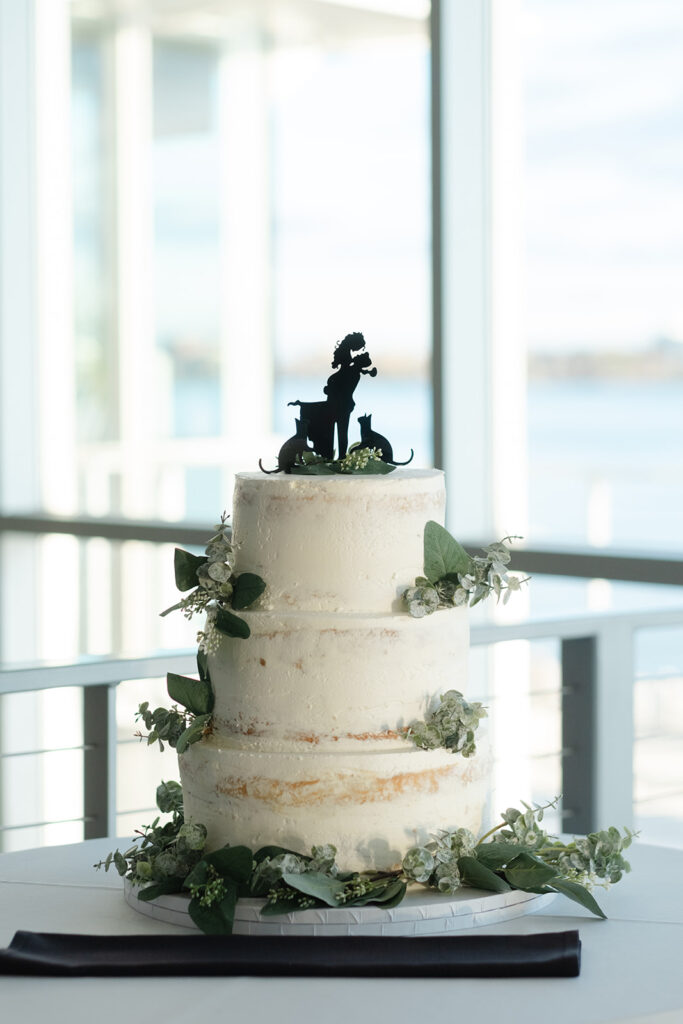 A simple, three-tier naked wedding cake with eucalyptus greenery, topped with a bride and pet silhouette cake topper.