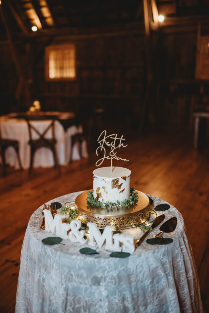 Rustic single-tier white wedding cake with gold leaf accents, on a gold stand, surrounded by “Mr. & Mrs.” décor.