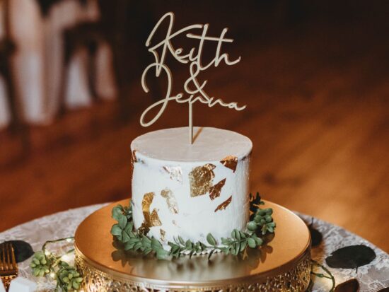 Close-up of a rustic single-tier white wedding cake with gold leaf detailing, greenery, and a personalized topper for the couple.