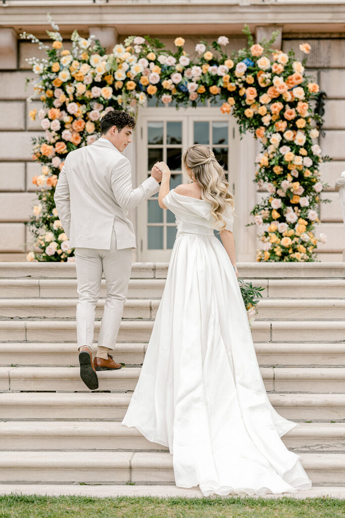 Couple walks up steps, bride’s gown flowing, surrounded by a flower arch with peach and pink blooms, in romantic setting.