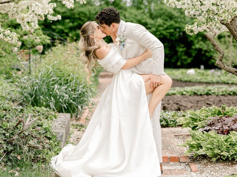 Bride and groom share a romantic kiss in a lush garden setting, with bride in elegant off-shoulder gown and long train.