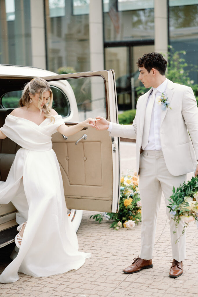 Bride steps out of vintage car in elegant off-shoulder gown, holding groom’s hand in a romantic outdoor setting.