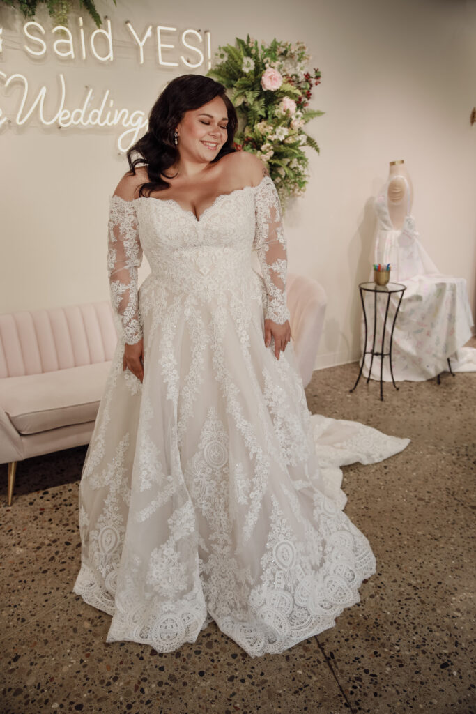 Radiant plus-size bride in a lace wedding gown, standing by a neon sign reading "I said YES!" at The Wedding Shoppe.