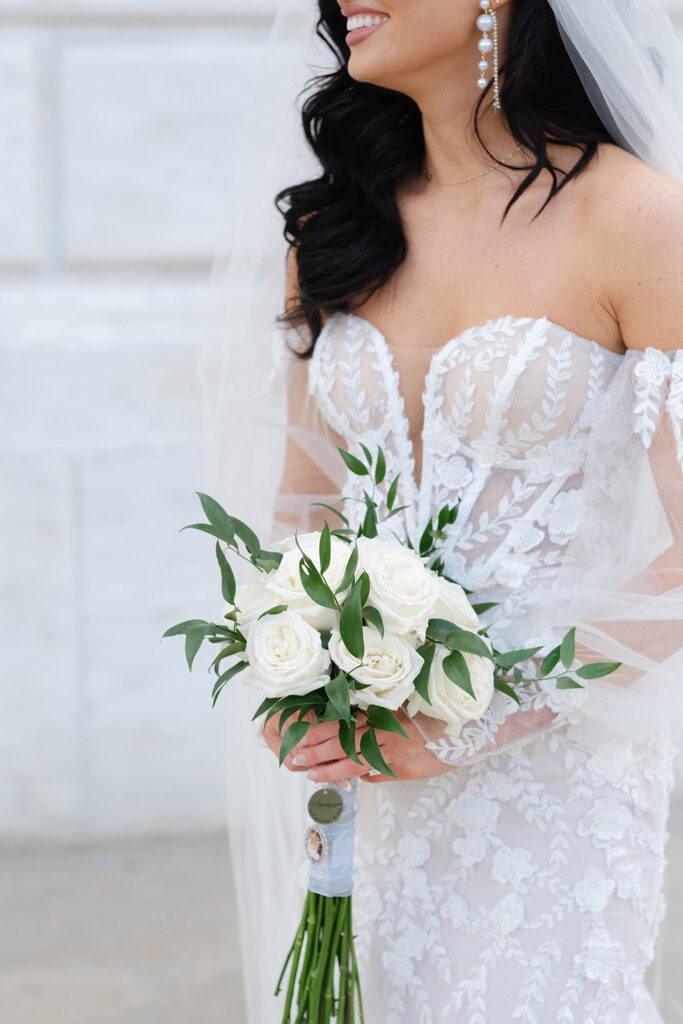 Bride in a lace dress holds a classic bouquet of white roses with greenery, highlighting her pearl drop earrings.