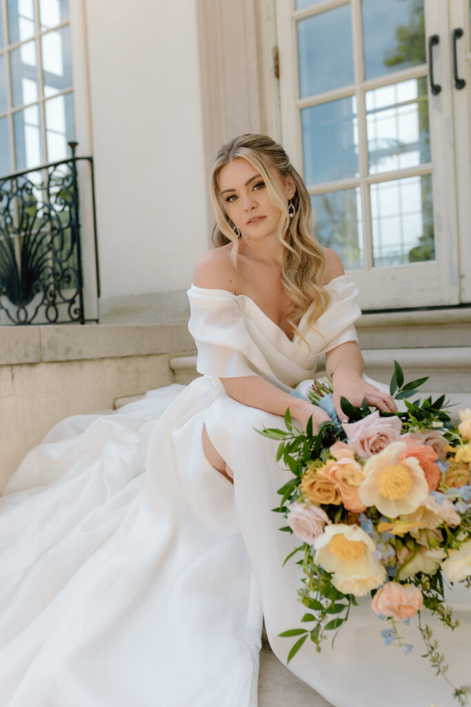 Bride sits gracefully on stone steps, holding a vibrant bouquet of yellow, peach, and pink flowers, gazing softly.