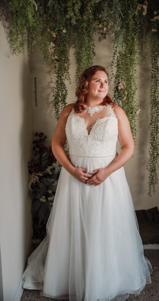 Bride in a sleeveless A-line gown with lace detailing, standing under greenery and floral décor at The Wedding Shoppe.