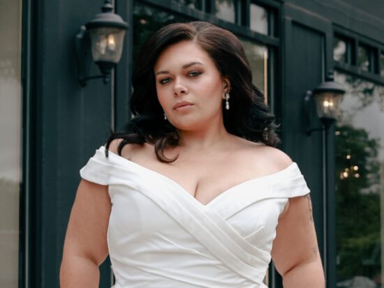 Confident plus-size bride in a sleek off-shoulder gown, posing outside The Wedding Shoppe with a determined expression.