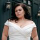 Confident plus-size bride in a sleek off-shoulder gown, posing outside The Wedding Shoppe with a determined expression.