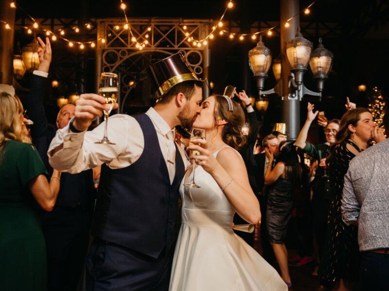 A couple kisses during a lively NYE wedding reception, surrounded by guests toasting with champagne under string lights.