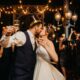 A couple kisses during a lively NYE wedding reception, surrounded by guests toasting with champagne under string lights.