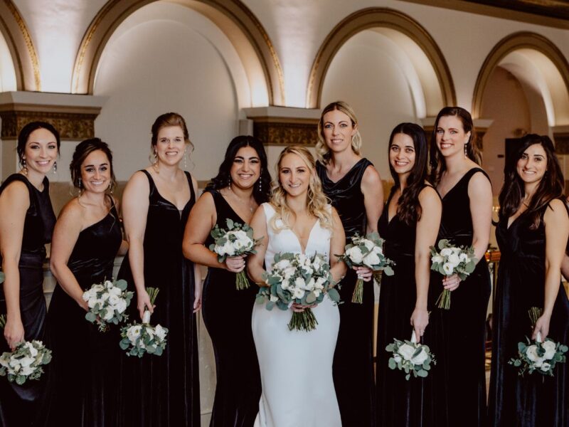 Bride with bridesmaids dressed in black dresses, holding white bouquets, posing in a luxurious venue with arches.