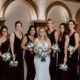 Bride with bridesmaids dressed in black dresses, holding white bouquets, posing in a luxurious venue with arches.