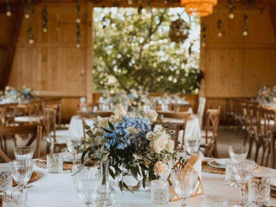A rustic wedding setup with tables featuring soft blue and white florals in a barn with warm natural wood tones.
