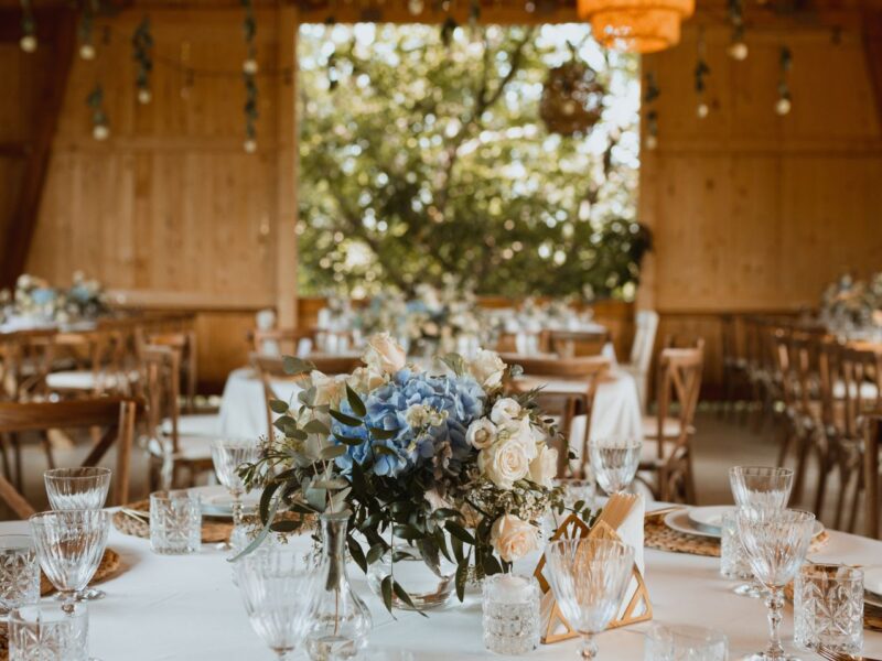 A rustic wedding setup with tables featuring soft blue and white florals in a barn with warm natural wood tones.