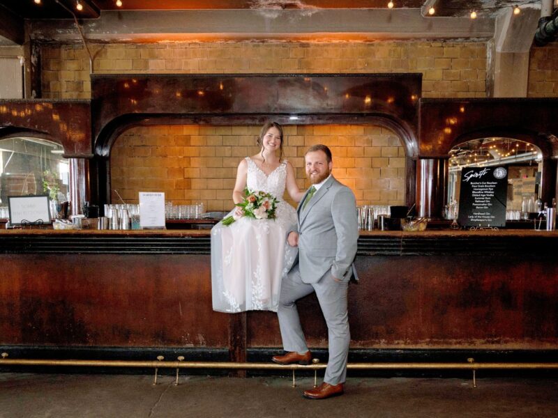 Bride perched on a vintage-style bar counter in a romantic lace dress, with her groom standing confidently beside her.