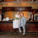 Bride perched on a vintage-style bar counter in a romantic lace dress, with her groom standing confidently beside her.