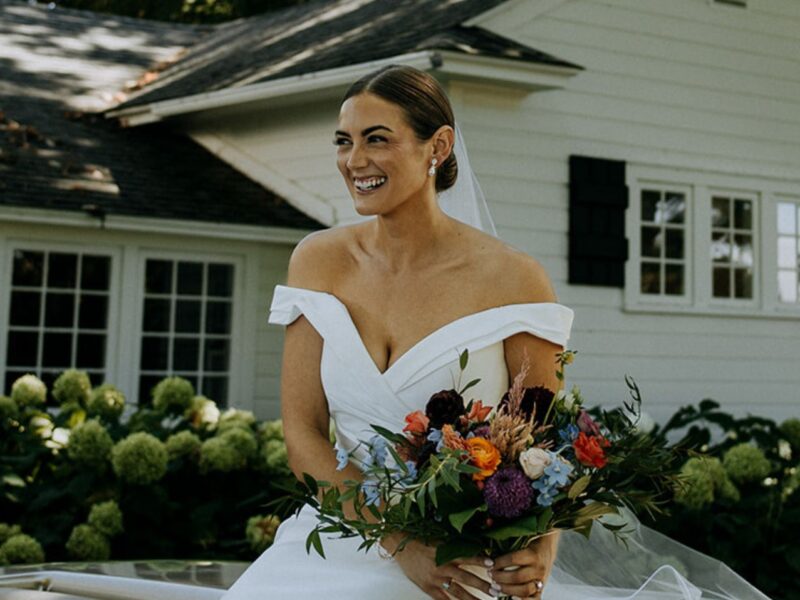 Bride in a modern off-shoulder gown holding a vibrant bouquet, seated outdoors near a white house with lush greenery.