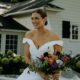 Bride in a modern off-shoulder gown holding a vibrant bouquet, seated outdoors near a white house with lush greenery.