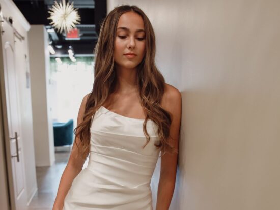 Close-up of a bride in a fitted off-the-shoulder satin gown with delicate ruching, standing against a chic boutique hallway.