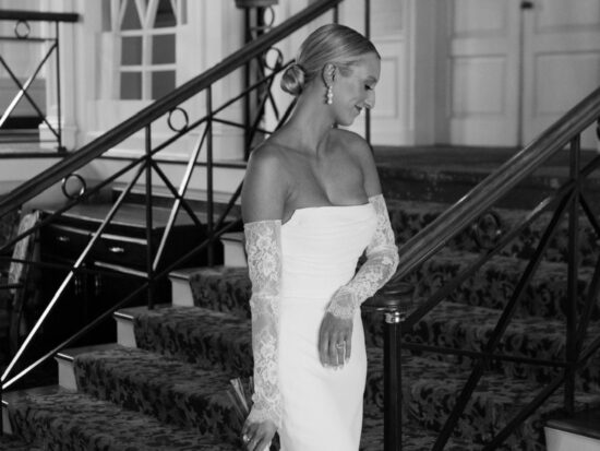 A black-and-white image captures a bride gracefully descending a staircase in a fitted wedding gown with lace sleeves.