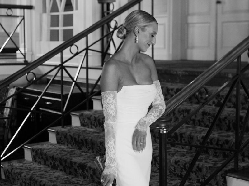 A black-and-white image captures a bride gracefully descending a staircase in a fitted wedding gown with lace sleeves.