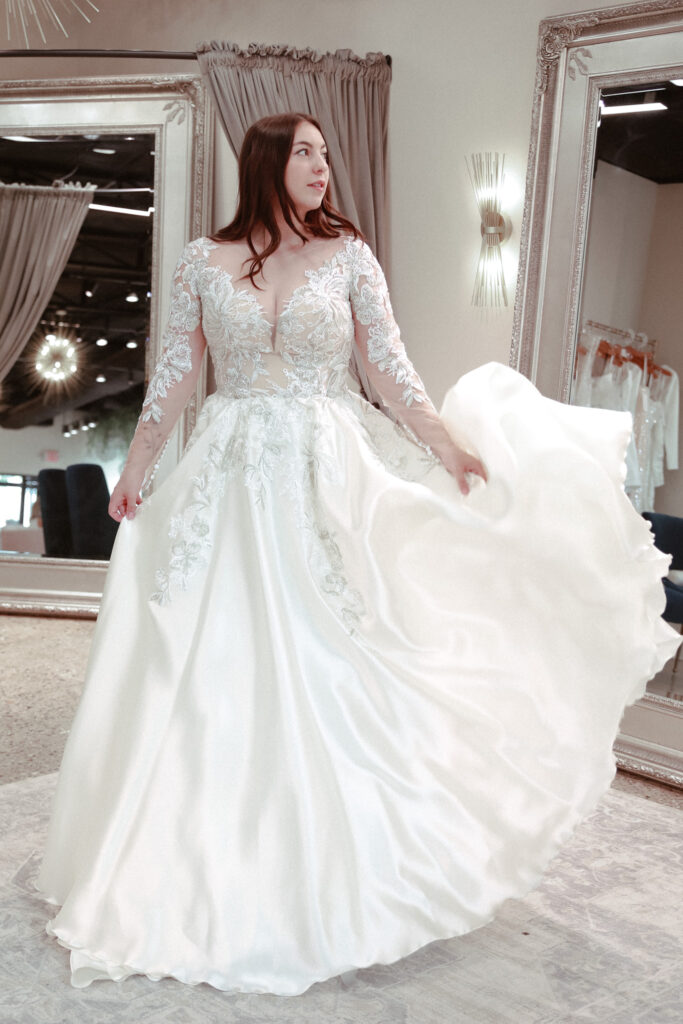 A bride elegantly twirls in a satin ball gown with long lace sleeves and a plunging neckline in a luxurious bridal boutique setting.