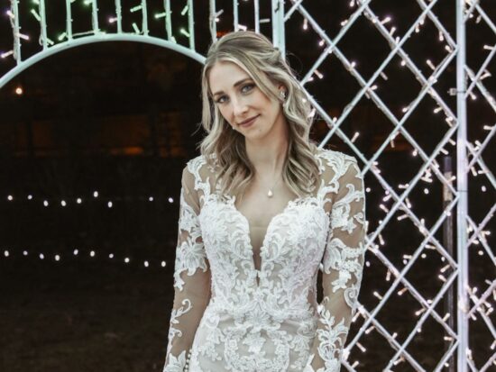 A bride smiles gracefully in a fitted lace gown with a sheer neckline, illuminated by twinkling fairy lights in a nighttime garden.
