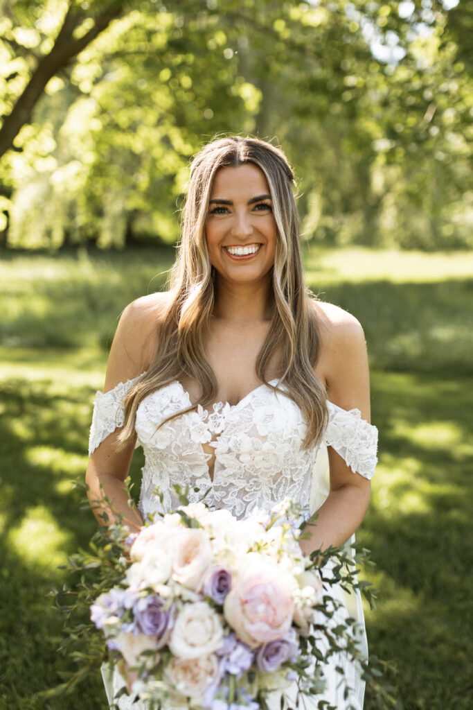 A beaming bride outdoors, dressed in an off-shoulder lace wedding gown, holding a bouquet of pastel pink, lavender, and cream roses.