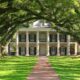 A grand Southern estate framed by moss-covered oak trees, leading to a picturesque entryway with white columns and lush greenery.