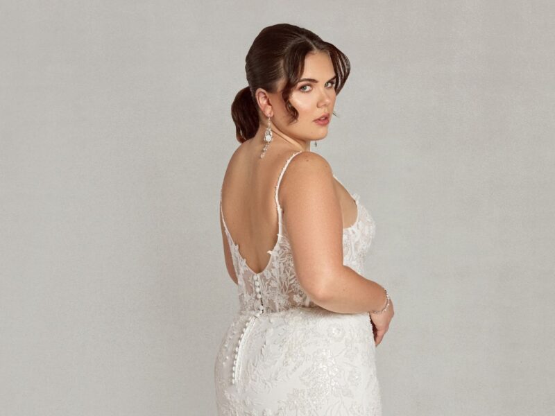 A bride wearing a floral lace fit-and-flare wedding gown with an open scoop back, standing against a neutral backdrop. The dress exudes vintage charm with modern sophistication.