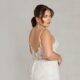 A bride wearing a floral lace fit-and-flare wedding gown with an open scoop back, standing against a neutral backdrop. The dress exudes vintage charm with modern sophistication.