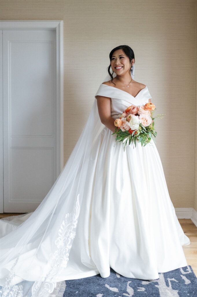 Elegant bride in an off-the-shoulder white gown with a flowing lace train, holding a bouquet of orange, peach, and white blooms indoors.