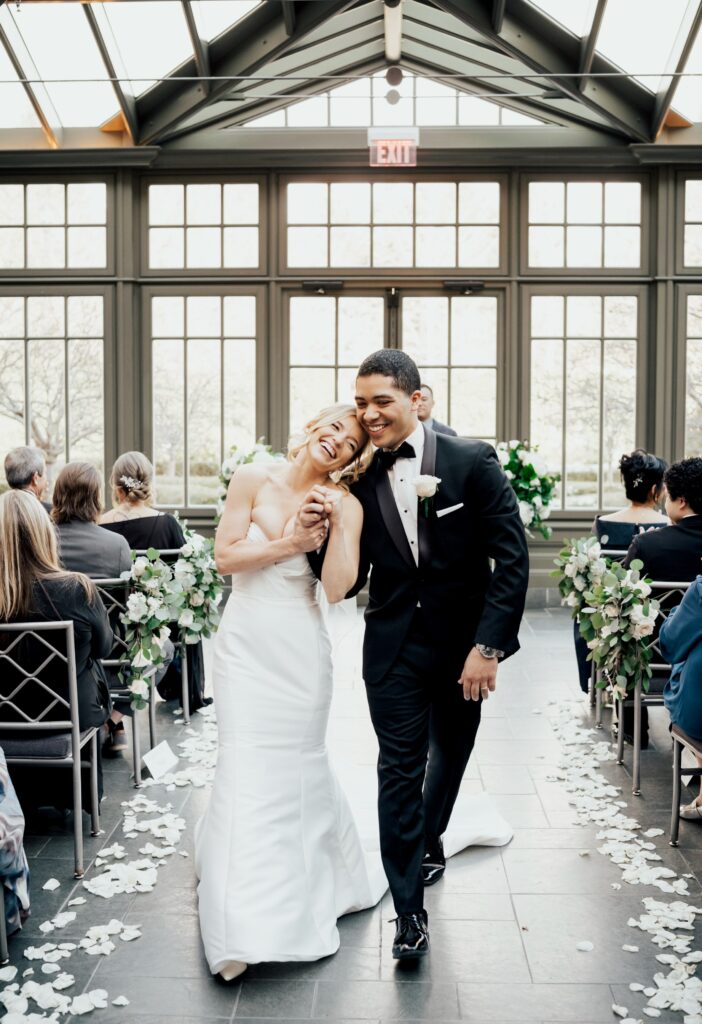 Joyful newlyweds holding hands and smiling as they walk down the aisle of a sunlit greenhouse with flower petals scattered on the floor.
