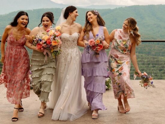 A bride and her bridesmaids laughing together, dressed in colorful floral and pastel dresses, against a scenic mountain backdrop.