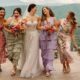 A bride and her bridesmaids laughing together, dressed in colorful floral and pastel dresses, against a scenic mountain backdrop.