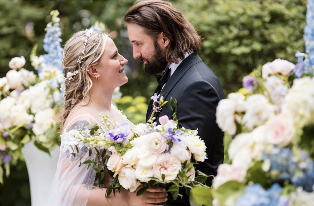 Bride and groom lovingly gazing into each other’s eyes, surrounded by lush florals in soft blue, ivory, and pastel pink hues.