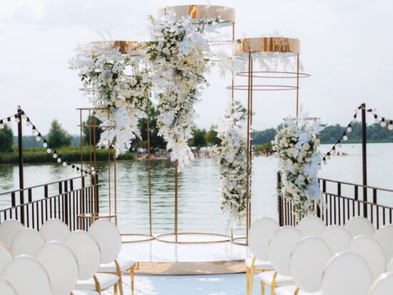 A stunning floral arch with white blossoms and gold accents, framed by rows of white chairs under string lights with a serene lake backdrop.