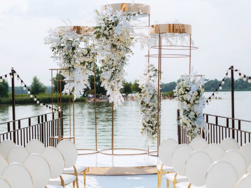 A stunning floral arch with white blossoms and gold accents, framed by rows of white chairs under string lights with a serene lake backdrop.