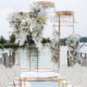 A stunning floral arch with white blossoms and gold accents, framed by rows of white chairs under string lights with a serene lake backdrop.