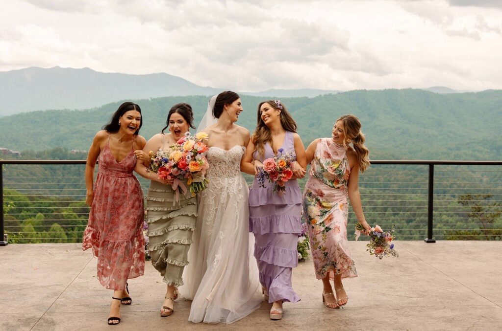 A bride and her bridesmaids laughing together, dressed in colorful floral and pastel dresses, against a scenic mountain backdrop.
