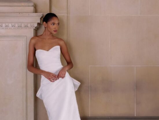 A bride in a strapless fit and flare wedding dress with subtle floral texture, leaning casually against an ornate stone column in a grand hallway.