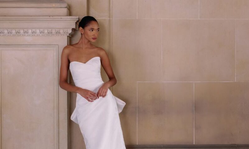 A bride in a strapless fit and flare wedding dress with subtle floral texture, leaning casually against an ornate stone column in a grand hallway.