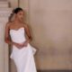 A bride in a strapless fit and flare wedding dress with subtle floral texture, leaning casually against an ornate stone column in a grand hallway.