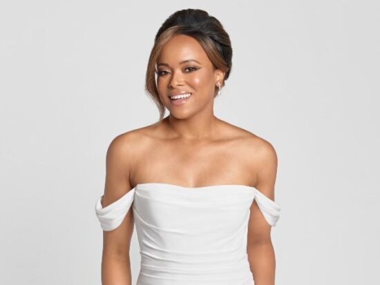 A smiling bride wears a white, off-the-shoulder wedding gown with a ruched bodice against a neutral background.