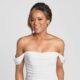 A smiling bride wears a white, off-the-shoulder wedding gown with a ruched bodice against a neutral background.
