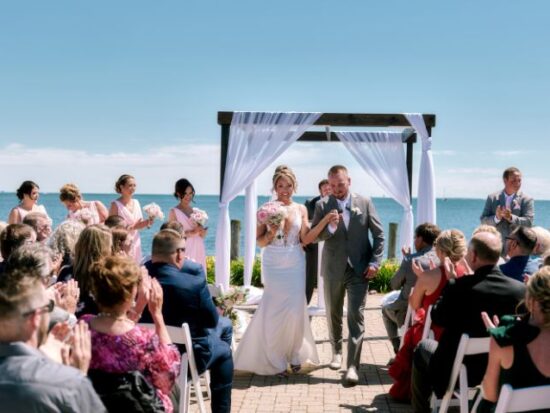 Bride in a fitted white gown with a long train smiles at the camera while holding hands with the groom in a navy suit, surrounded by garden greenery.